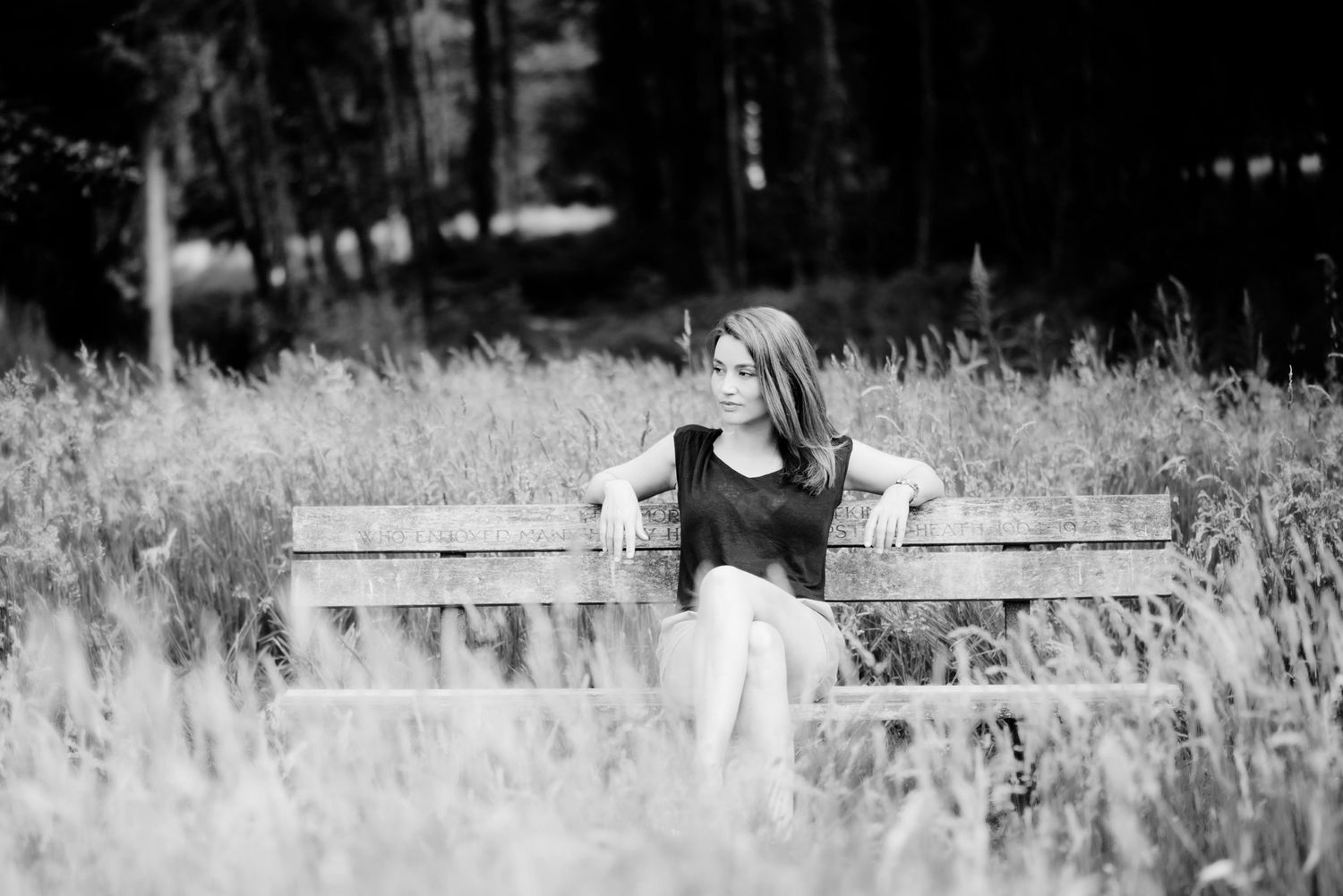 Young woman in black outfit sitting on a wooden bench surrounded by tall grass and trees in a black and white photo.