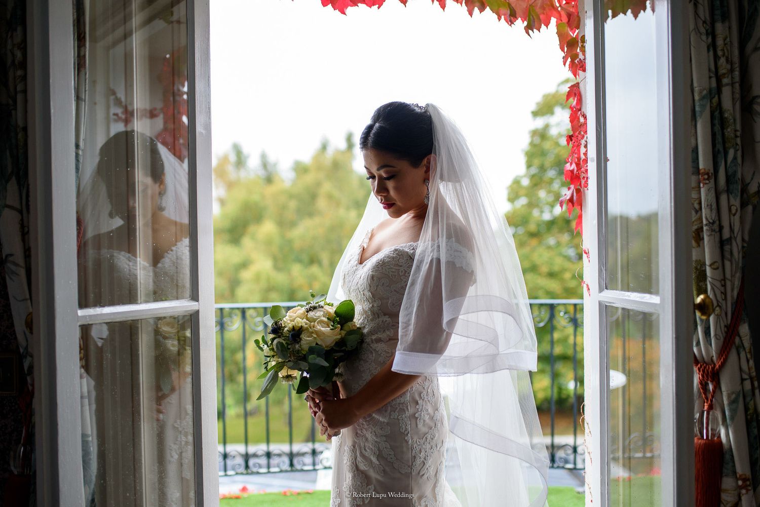 Bride in white lace gown and veil holds bouquet by open French doors with autumn trees in background.