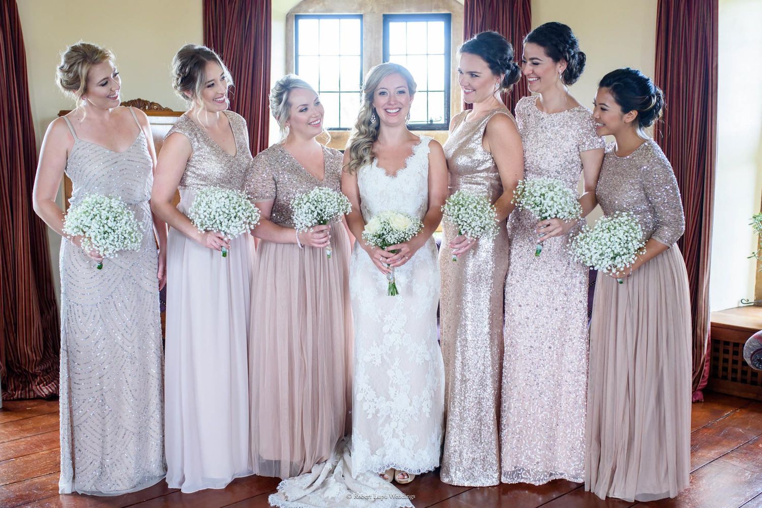 Bride in white lace gown poses with six bridesmaids in blush and gold sequin dresses holding white baby's breath bouquets.