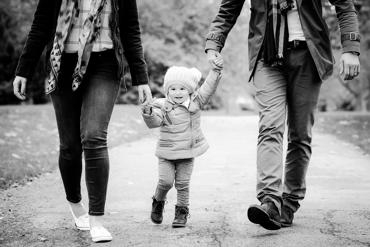 Black and white photo of parents holding hands with toddler walking on a path outdoors.