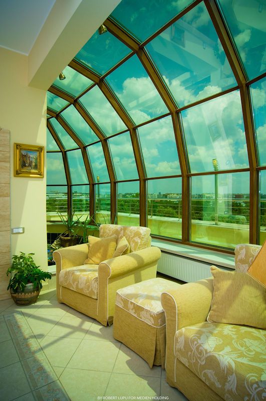 Sunroom with arched glass ceiling, yellow floral sofas, tile floors, and panoramic city views under a blue sky.