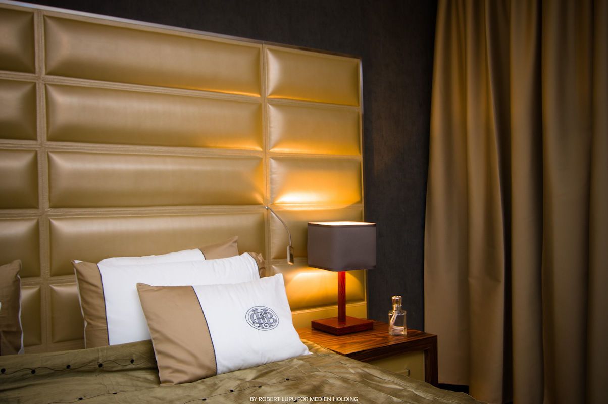 Luxury hotel bedroom with gold tufted headboard, decorative pillows, bedside lamp, and elegant brown curtains.