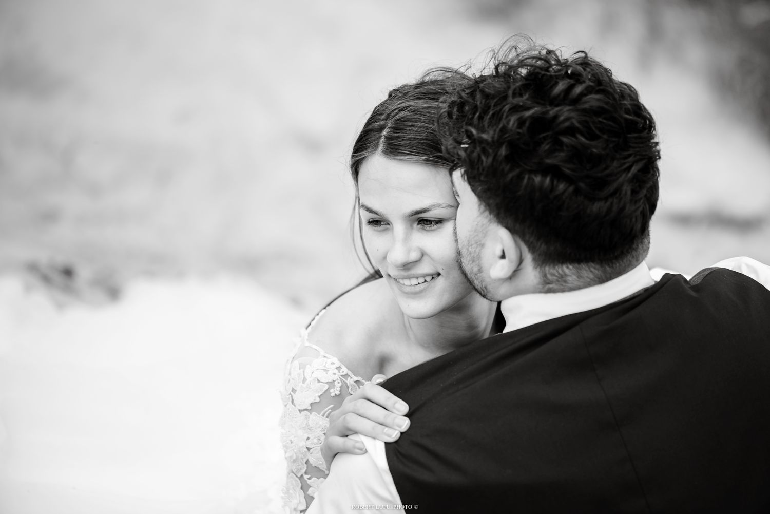 Radiant bride in lace gown smiles while embracing groom in black suit in a tender black and white wedding portrait.