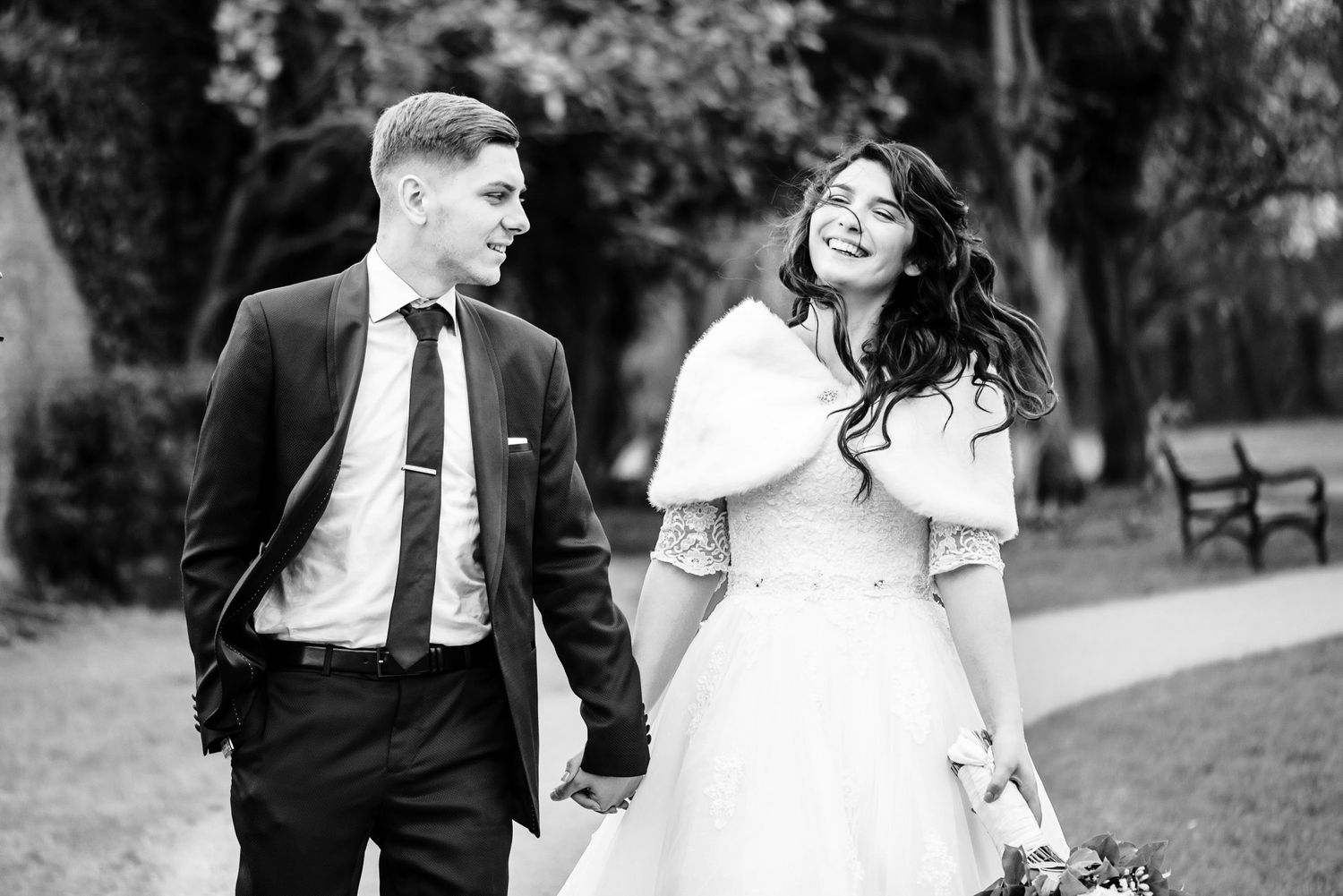 Black and white photo of a smiling bride and groom holding hands while walking in a park on their wedding day.