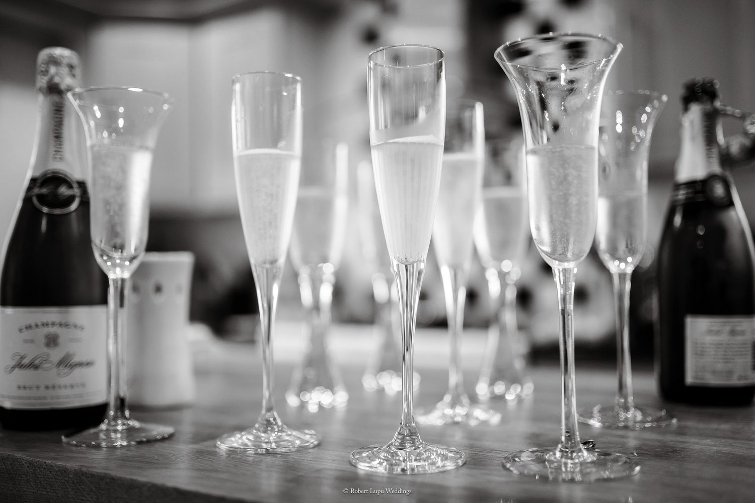 Black and white photo of champagne flutes filled with sparkling wine beside bottles on a bar.