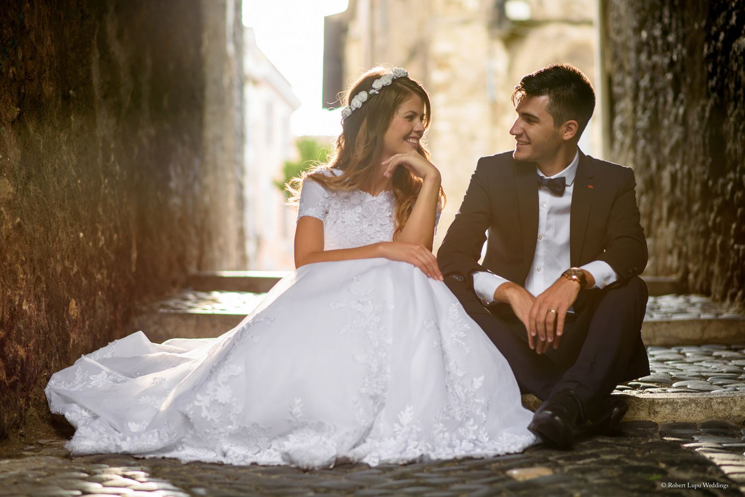 A bride in a white ball gown and groom in a black tuxedo sit on cobblestone steps sharing a romantic moment.