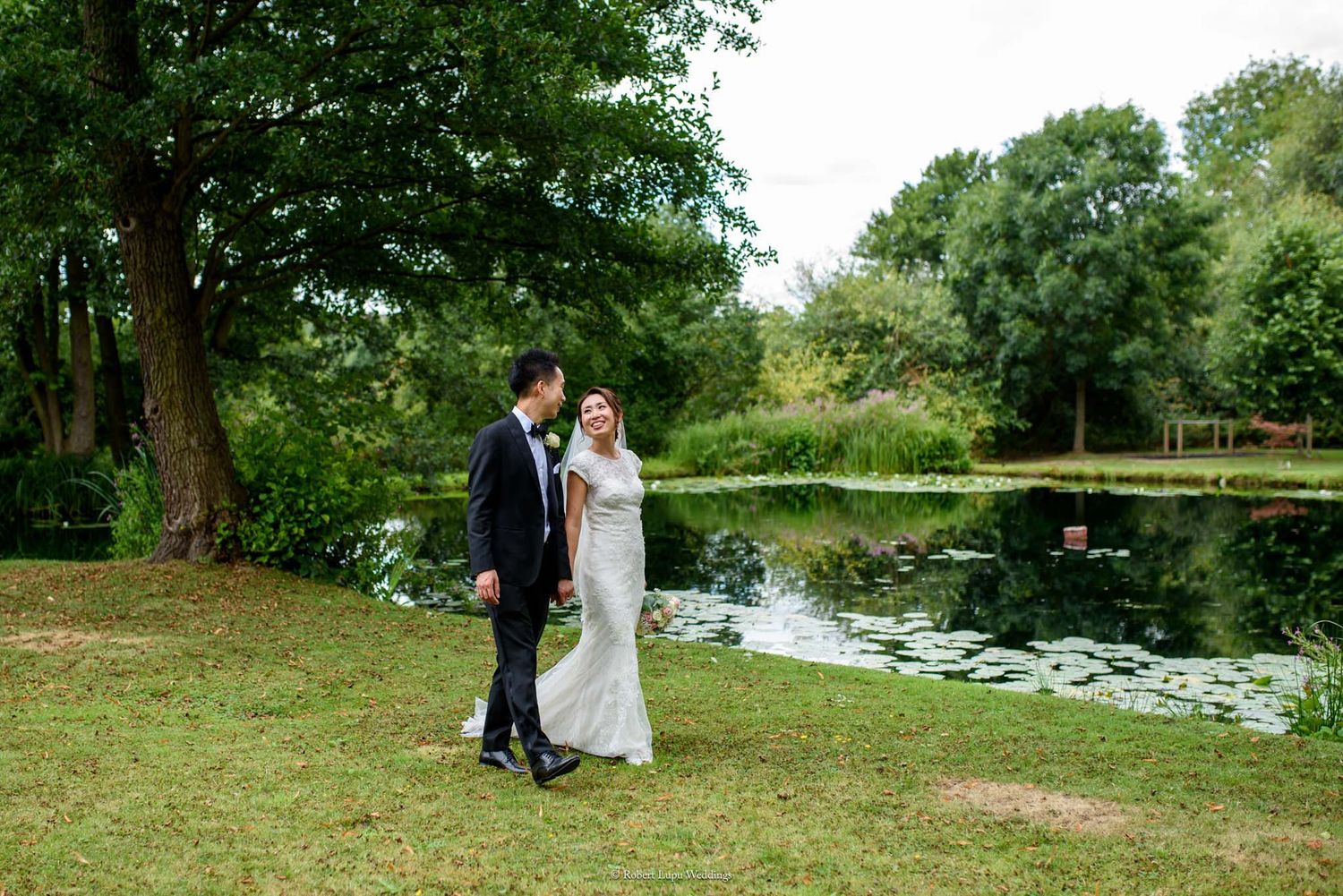 Bride and groom stroll beside a serene pond surrounded by lush greenery on their wedding day.