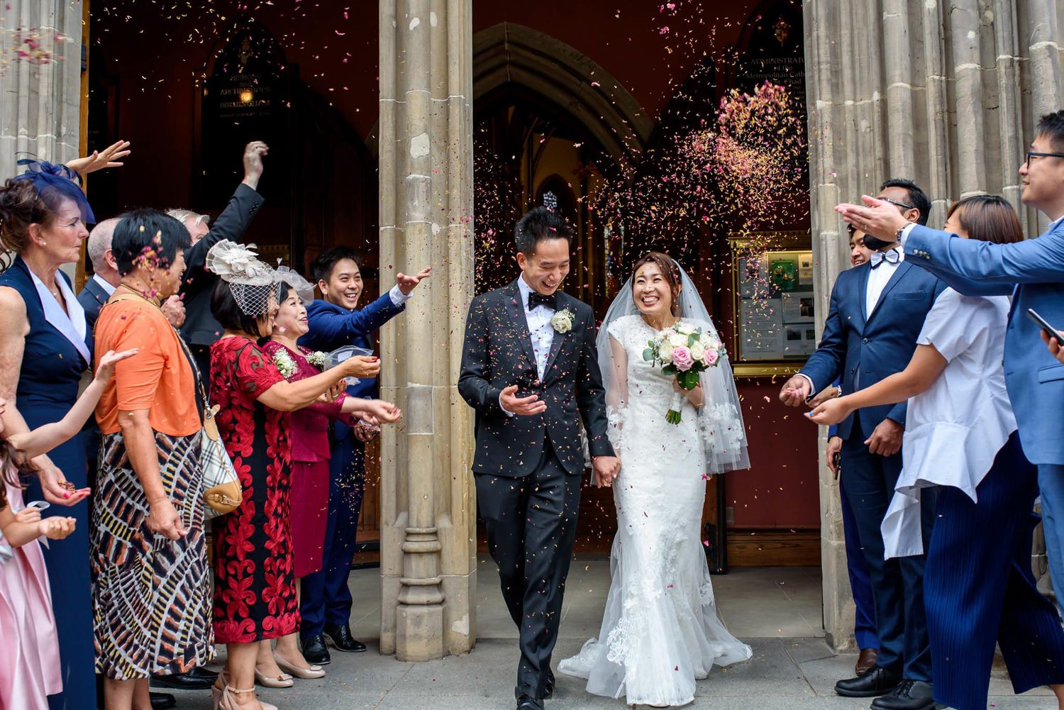 Newlywed Asian couple exits church as guests toss confetti in joyful wedding celebration.