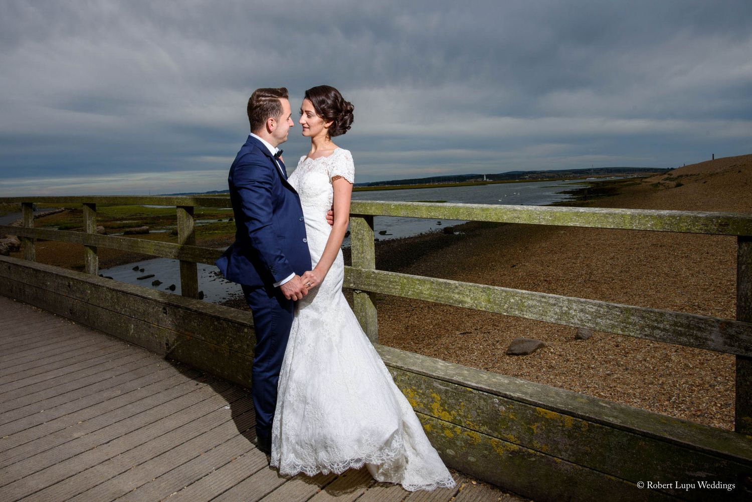 Bride and groom share a romantic moment on a coastal boardwalk under dramatic stormy skies at their seaside wedding.