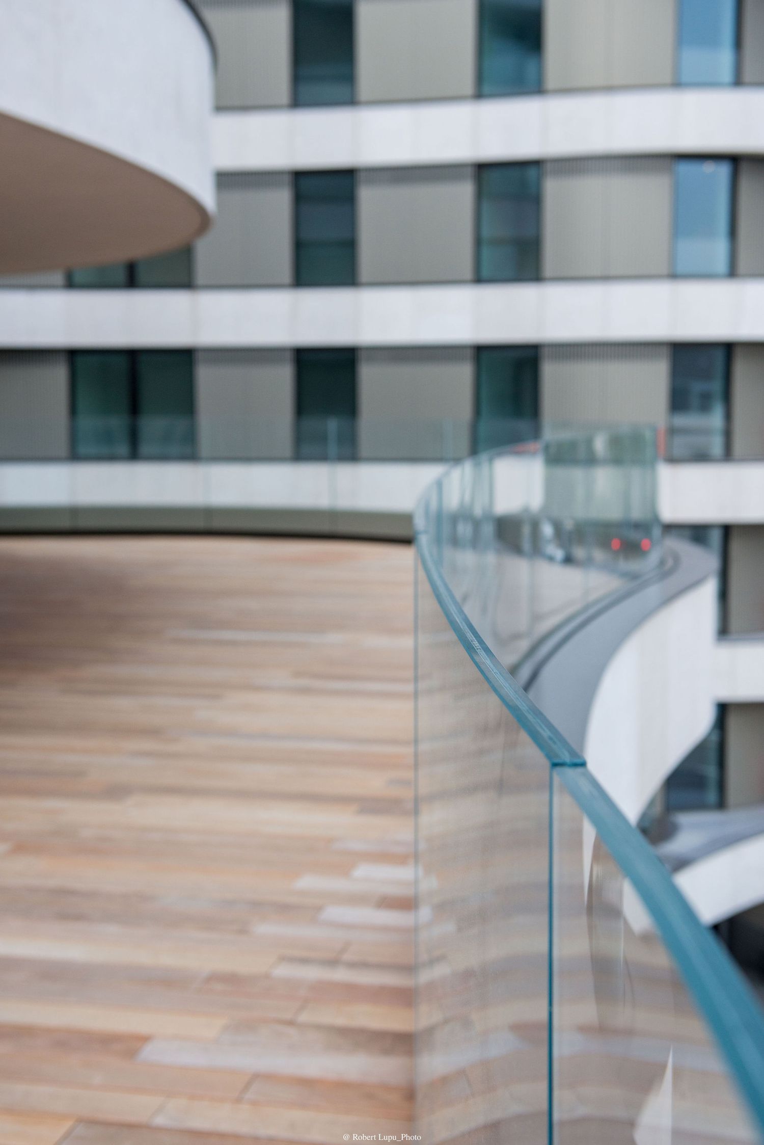 Modern curved glass balcony railing with wooden deck overlooking a contemporary white multi-story building facade.