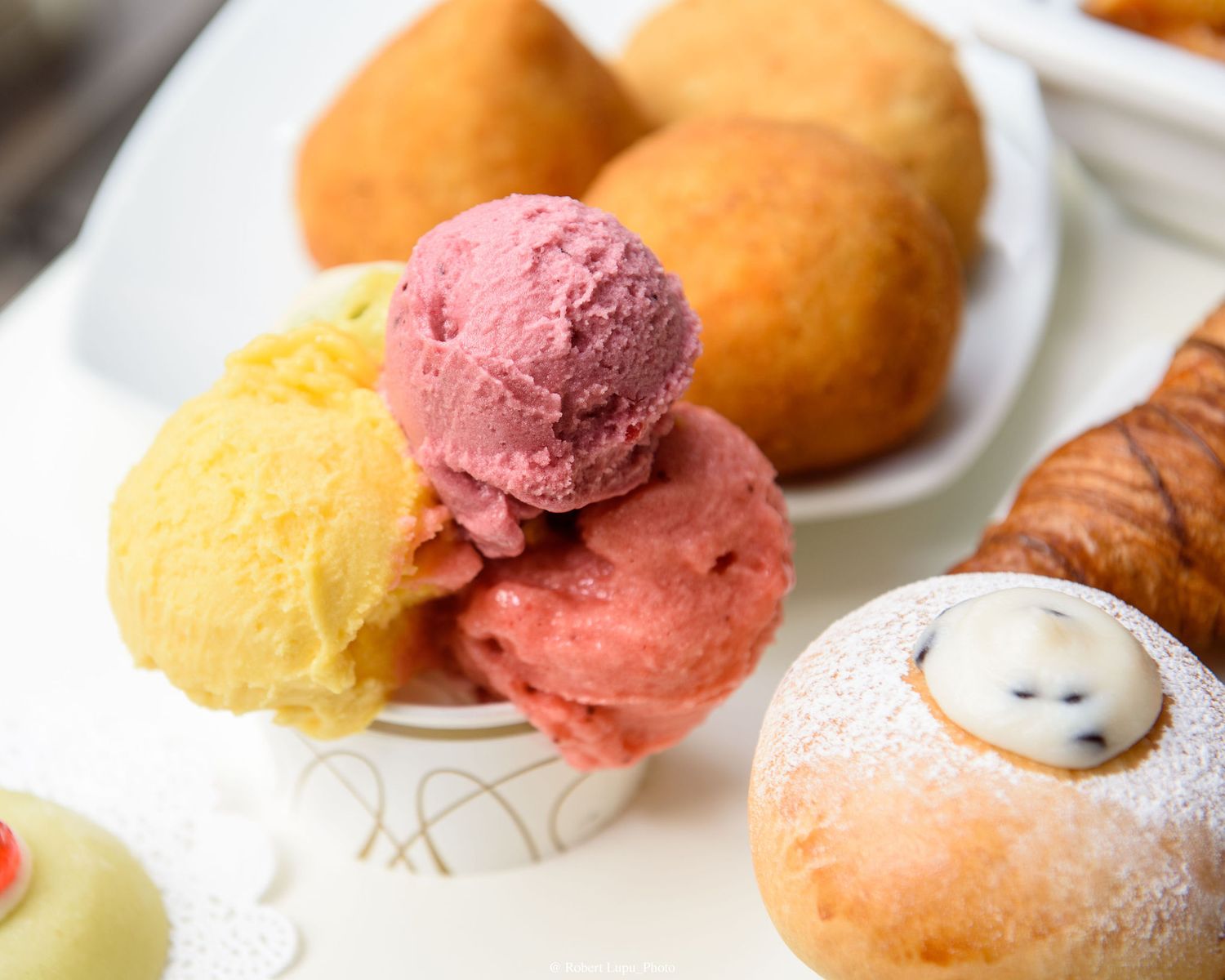Colorful scoops of gelato in a cup beside fried pastries and a powdered sugar-dusted bun.