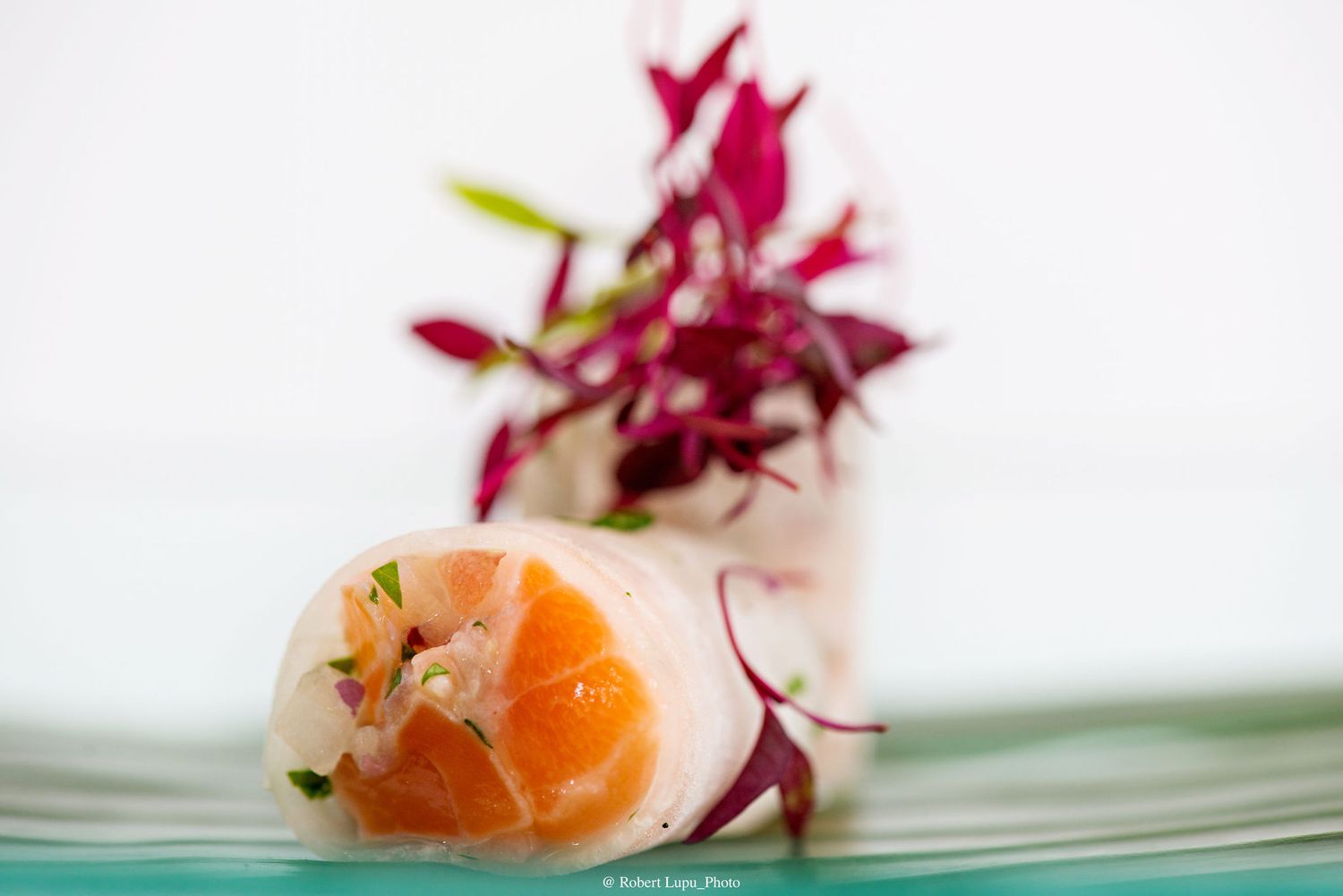 Elegant spring roll with fresh salmon and shrimp, garnished with vibrant red flowers on a teal plate.