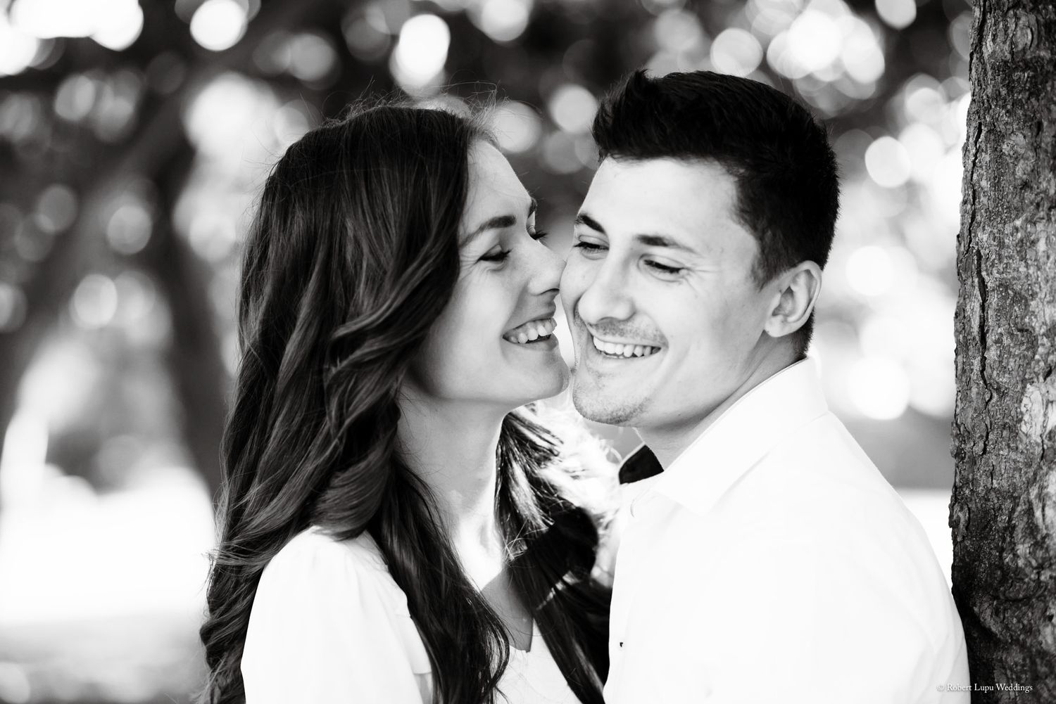 Black and white photo of a smiling couple, woman kissing man on cheek near a tree outdoors.