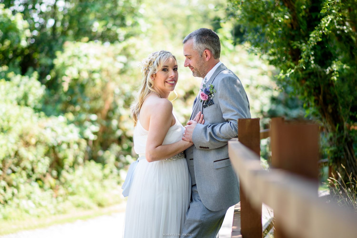 Smiling bride and groom in gray suit embrace outdoors near wooden fence surrounded by lush green garden on wedding day.