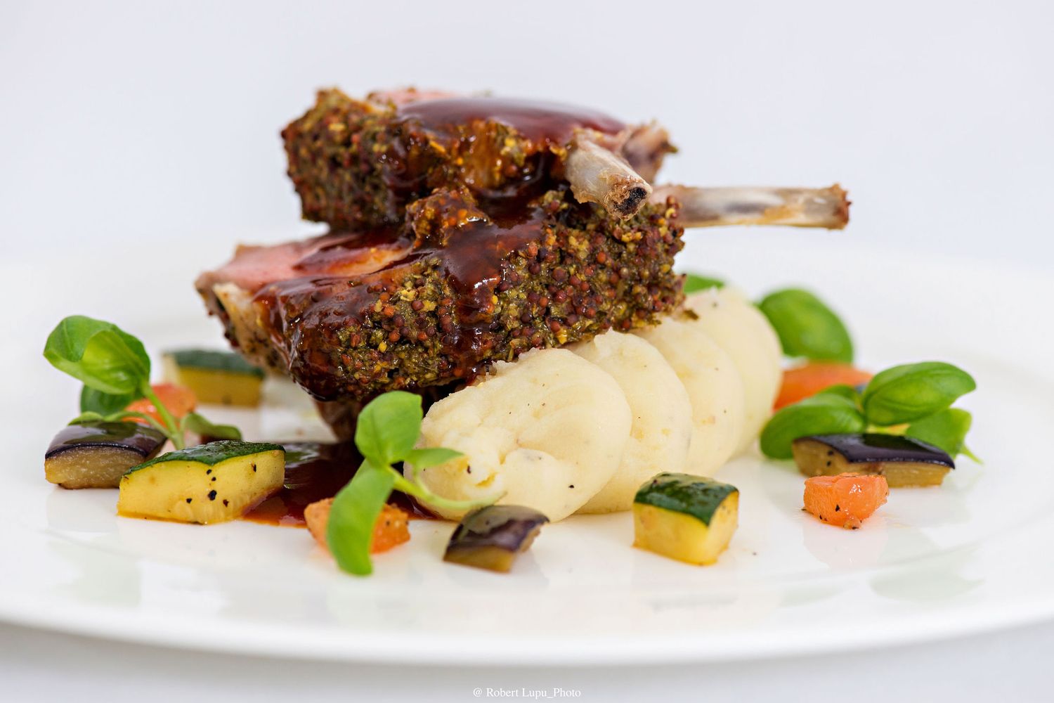 Herb-crusted lamb chops served with creamy mashed potatoes, grilled zucchini, carrots, and fresh basil on a white plate.