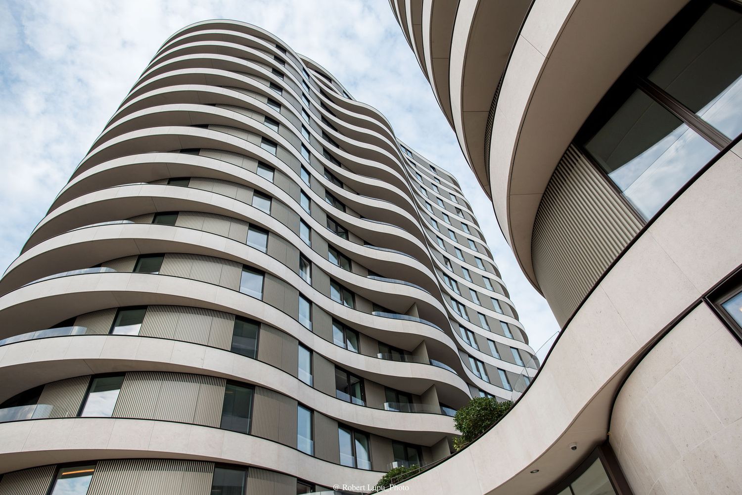 Modern curved high-rise apartment building with white balconies against a cloudy sky, viewed from below.