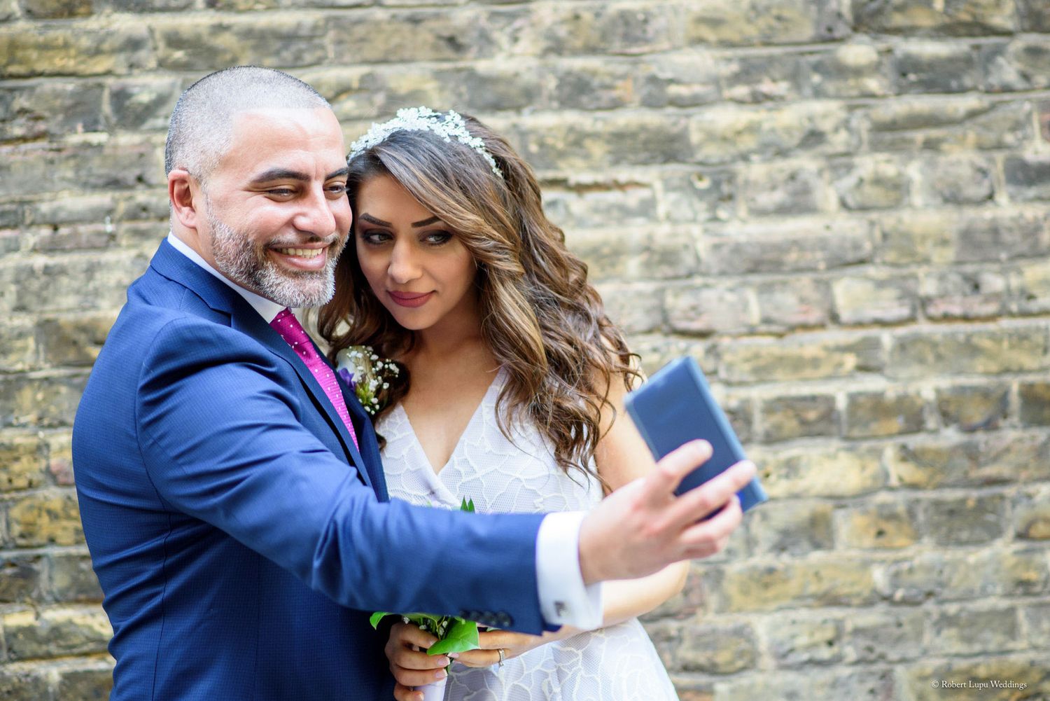Bride and groom taking a selfie together against a brick wall.
