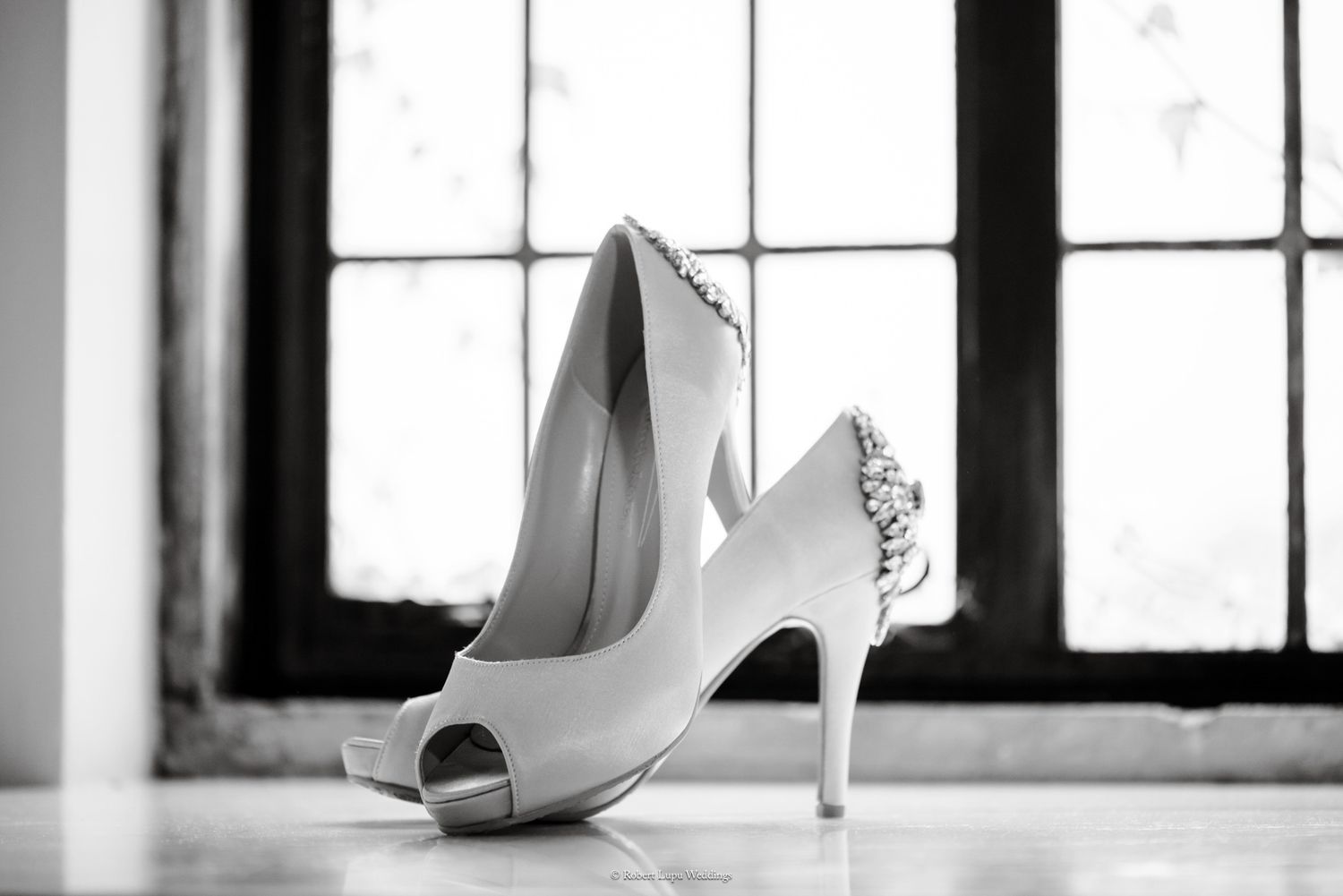 Elegant white bridal heels with jeweled detail displayed near window in black and white photo.