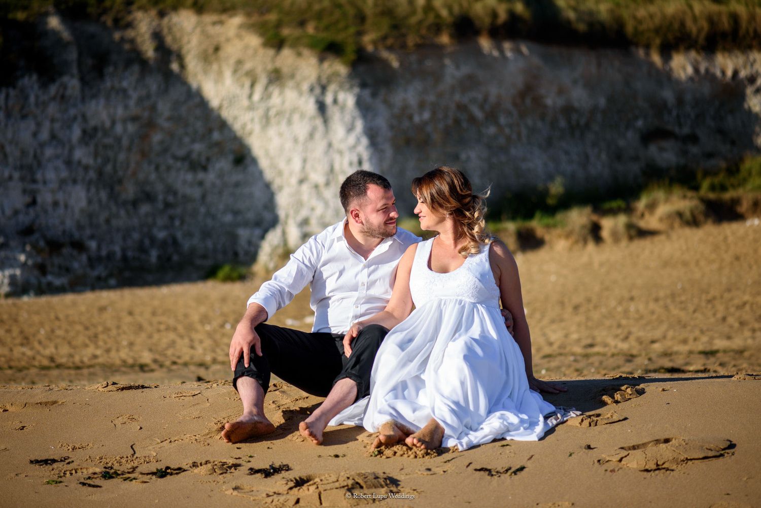 Couple in white outfits sitting on sandy beach with white cliffs, sharing a romantic moment.