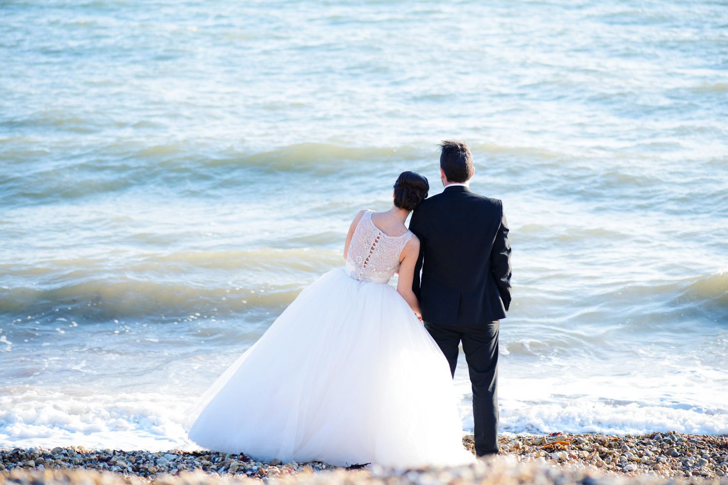 Bride and groom standing together on a rocky beach shore, gazing at ocean waves on their wedding day.