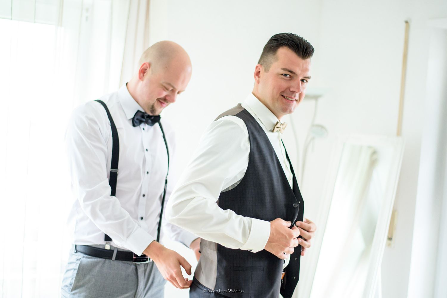 Two grooms preparing for wedding, one adjusting vest while other wears suspenders and bow tie in bright room.