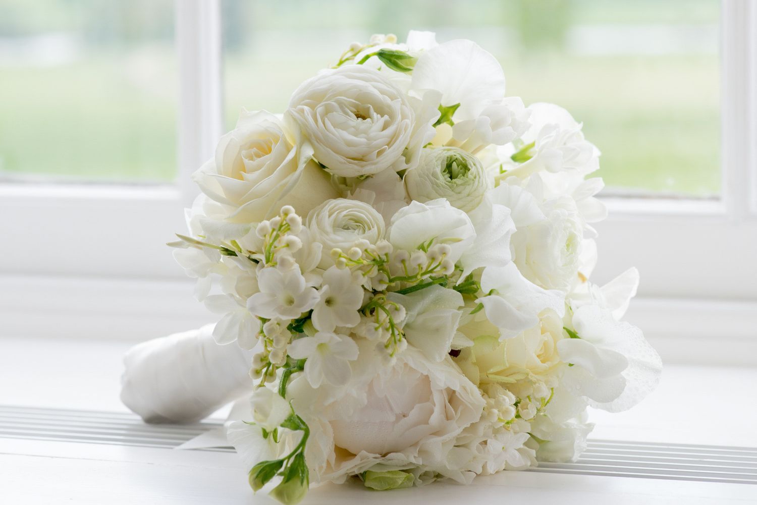 Elegant white bridal bouquet of roses, peonies, and freesia on a window sill in natural light.