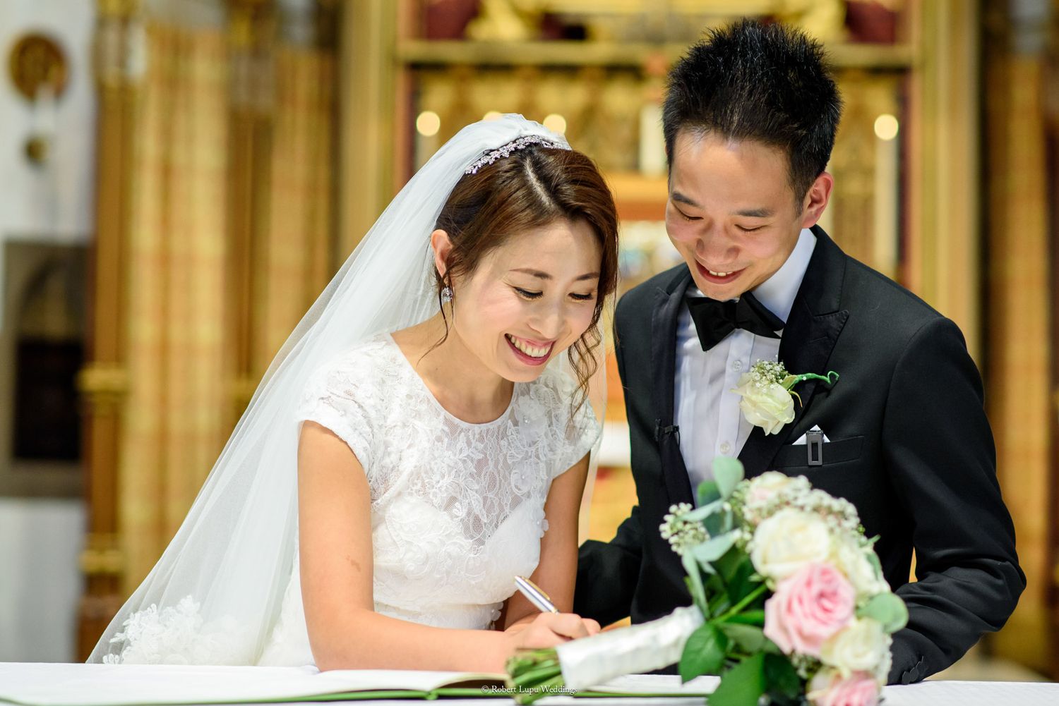 Smiling Asian bride and groom signing wedding register in elegant venue, bride wearing lace dress and veil.