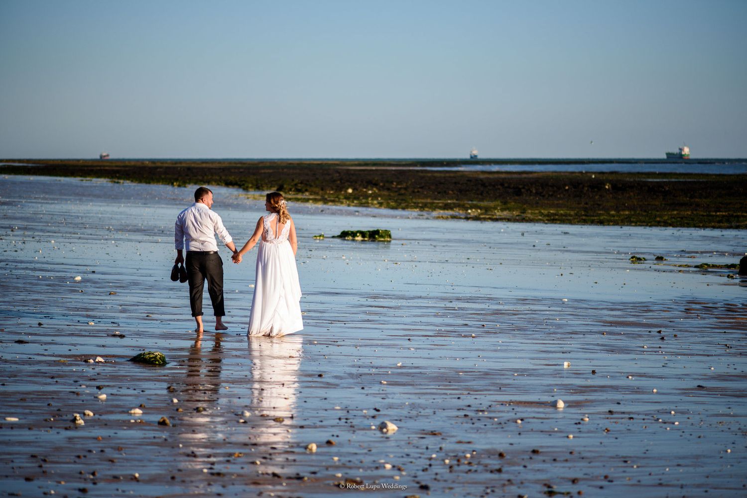 Newlywed couple holding hands walking on a reflective beach at sunset in wedding attire.