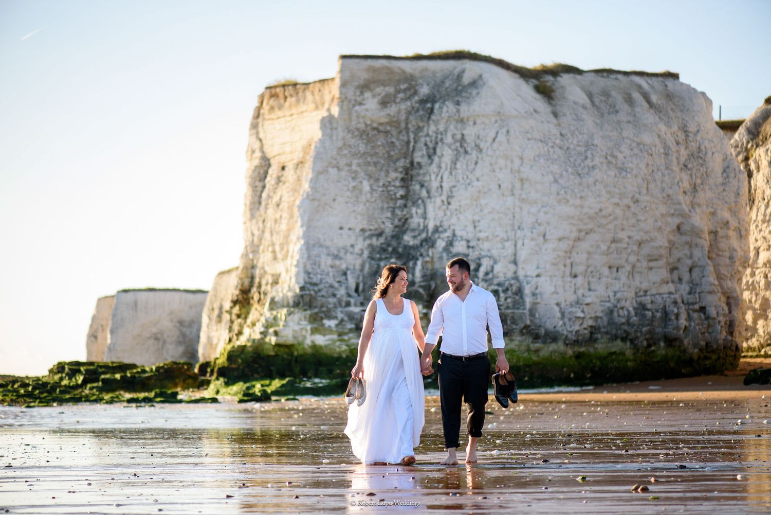 Couple in wedding attire walking hand in hand on a beach with white chalk cliffs in the background.