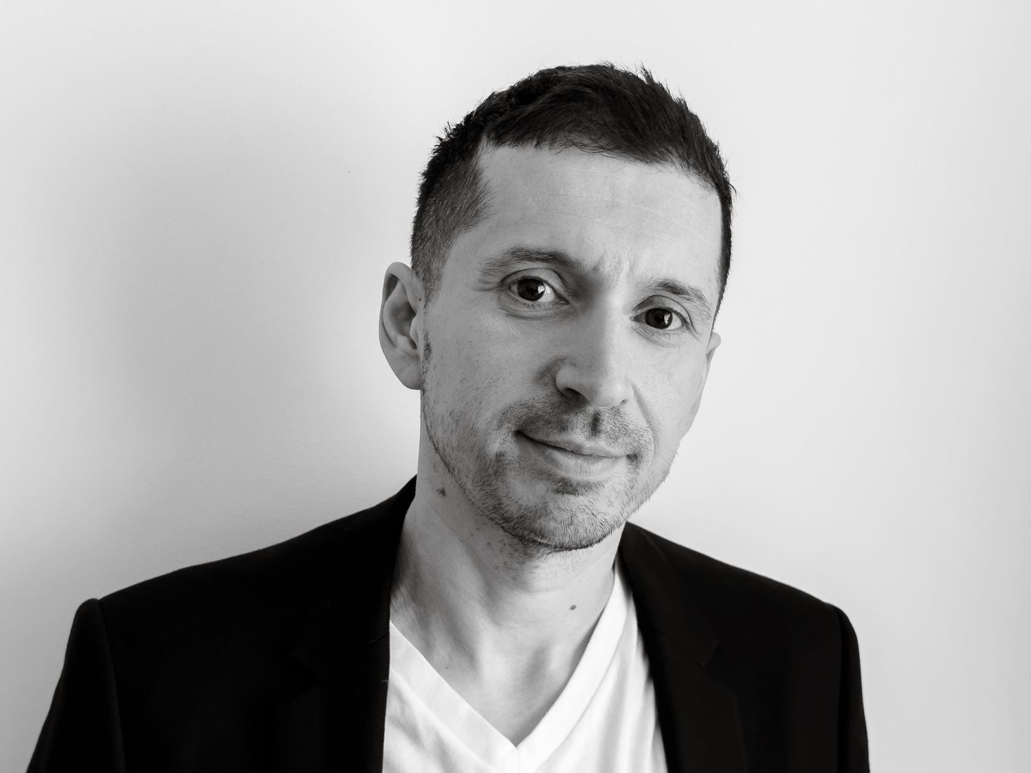 Black and white professional headshot of a man in a blazer and white V-neck shirt smiling confidently.