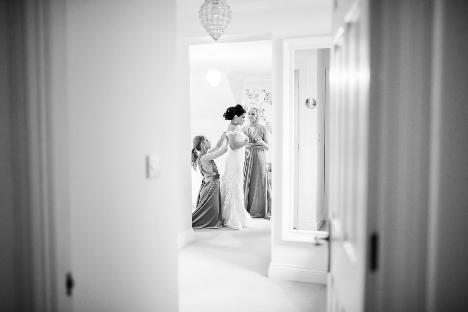 Black and white photo of bridesmaids helping a bride prepare in a bright hallway.