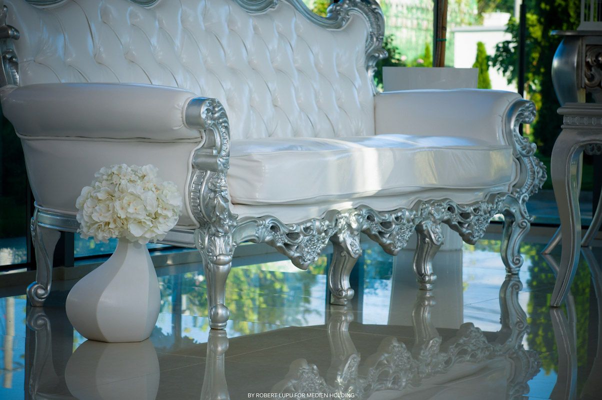Elegant white tufted baroque sofa with silver ornate frame on reflective floor beside white hydrangea vase.