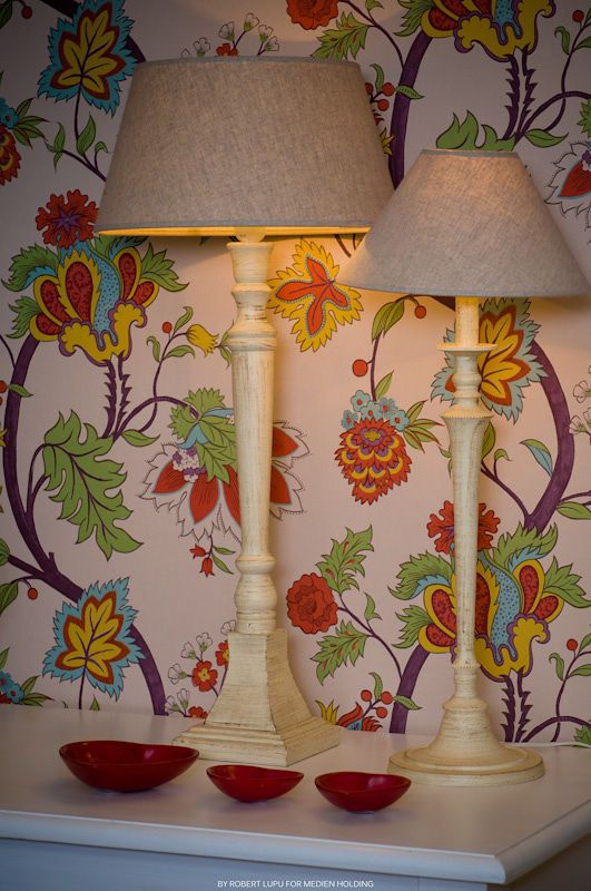 Two cream vintage-style table lamps on a white console against vibrant floral wallpaper with red bowls.