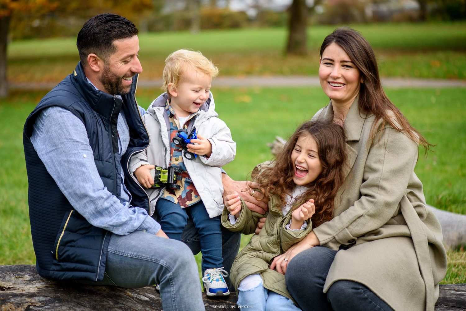 Happy family of four sitting outdoors in a park, parents smiling with young children on an autumn day.