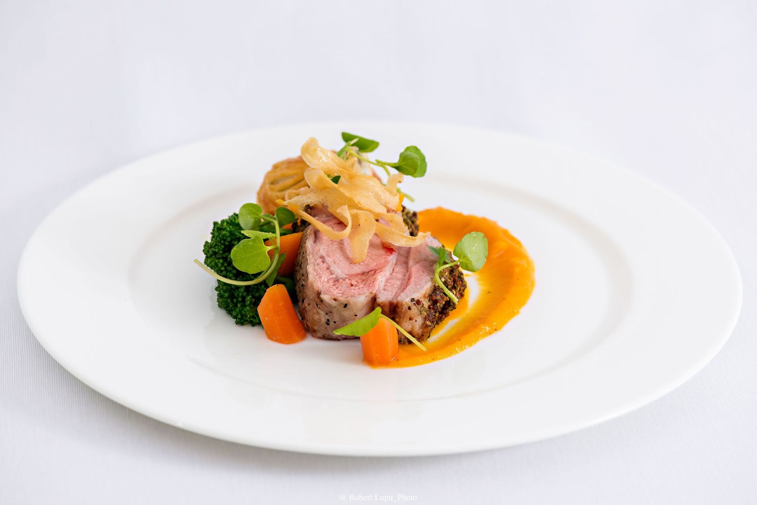Elegant plated dish featuring sliced rosy lamb, vibrant broccoli, glazed carrots, crispy onions, and orange sauce on white plate.