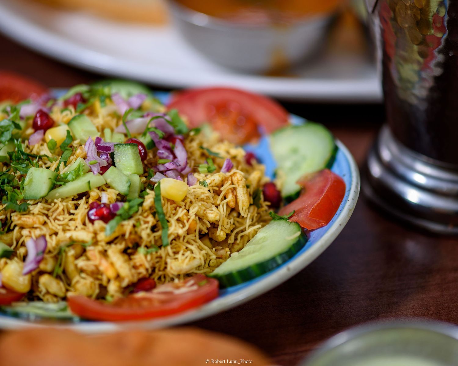 Colorful Indian chaat dish with sev, pomegranate, red onion, tomatoes, and cucumbers on a decorative plate.