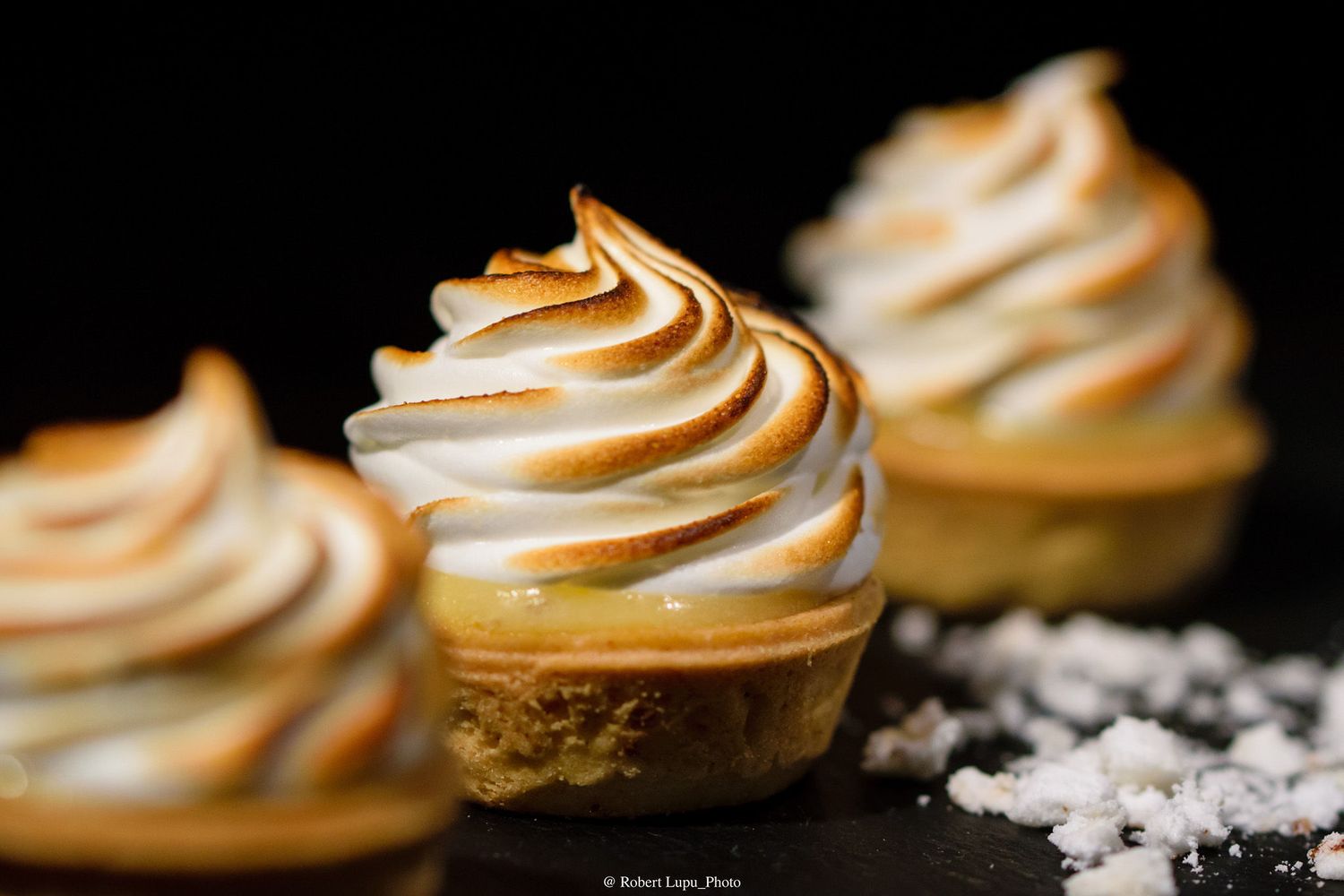 Mini lemon meringue tarts with golden-toasted swirled meringue peaks on a dark slate surface with sea salt flakes.