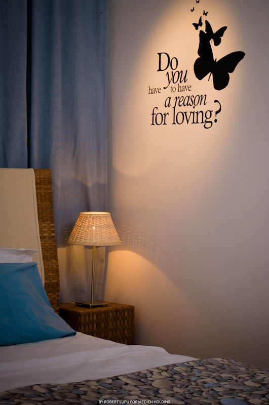 Cozy bedroom with wicker headboard, glowing table lamp, and butterfly wall decal reading Do you have to have a reason for loving.