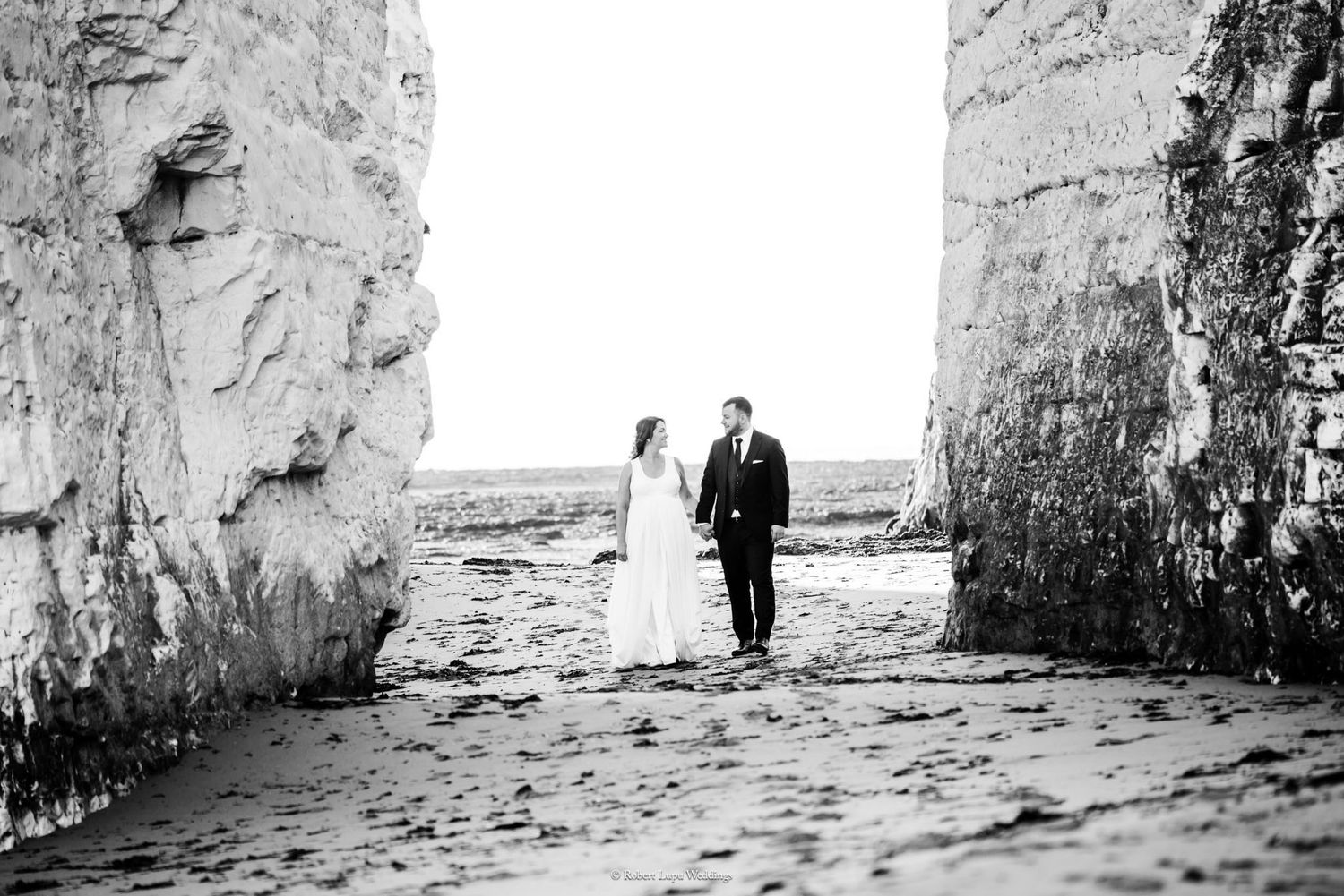 Bride and groom walking hand in hand between towering white chalk cliffs on a beach.