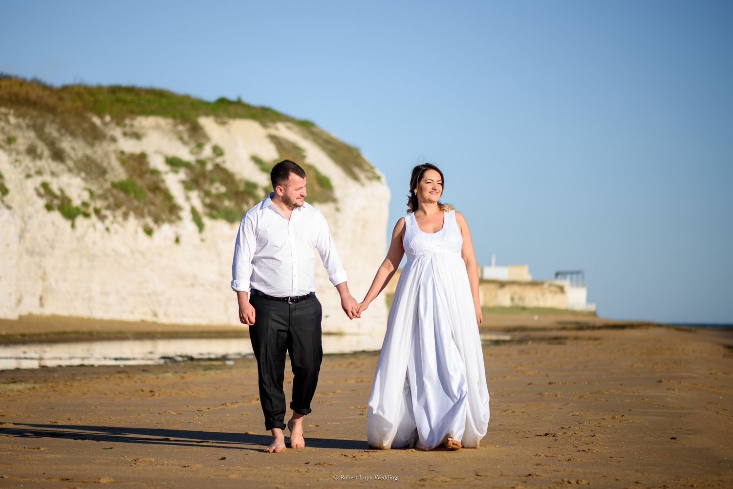 Couple in wedding attire walking hand in hand on a sunny beach with white cliffs in the background.