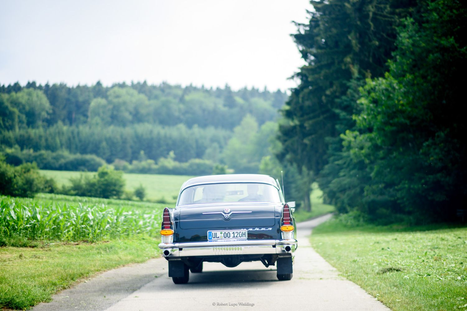 Vintage blue classic car driving down a narrow rural road surrounded by lush green fields and forest trees.