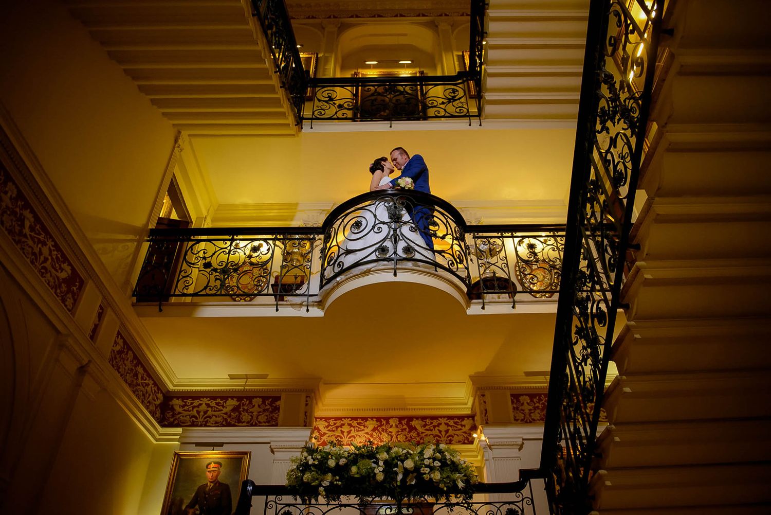 Bride and groom share a romantic kiss on an ornate balcony with elegant iron railings in a grand wedding venue.