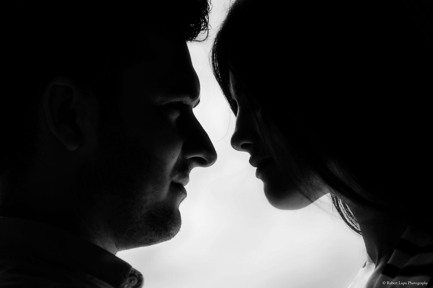 Intimate black and white silhouette of couple facing each other close together against bright sky.
