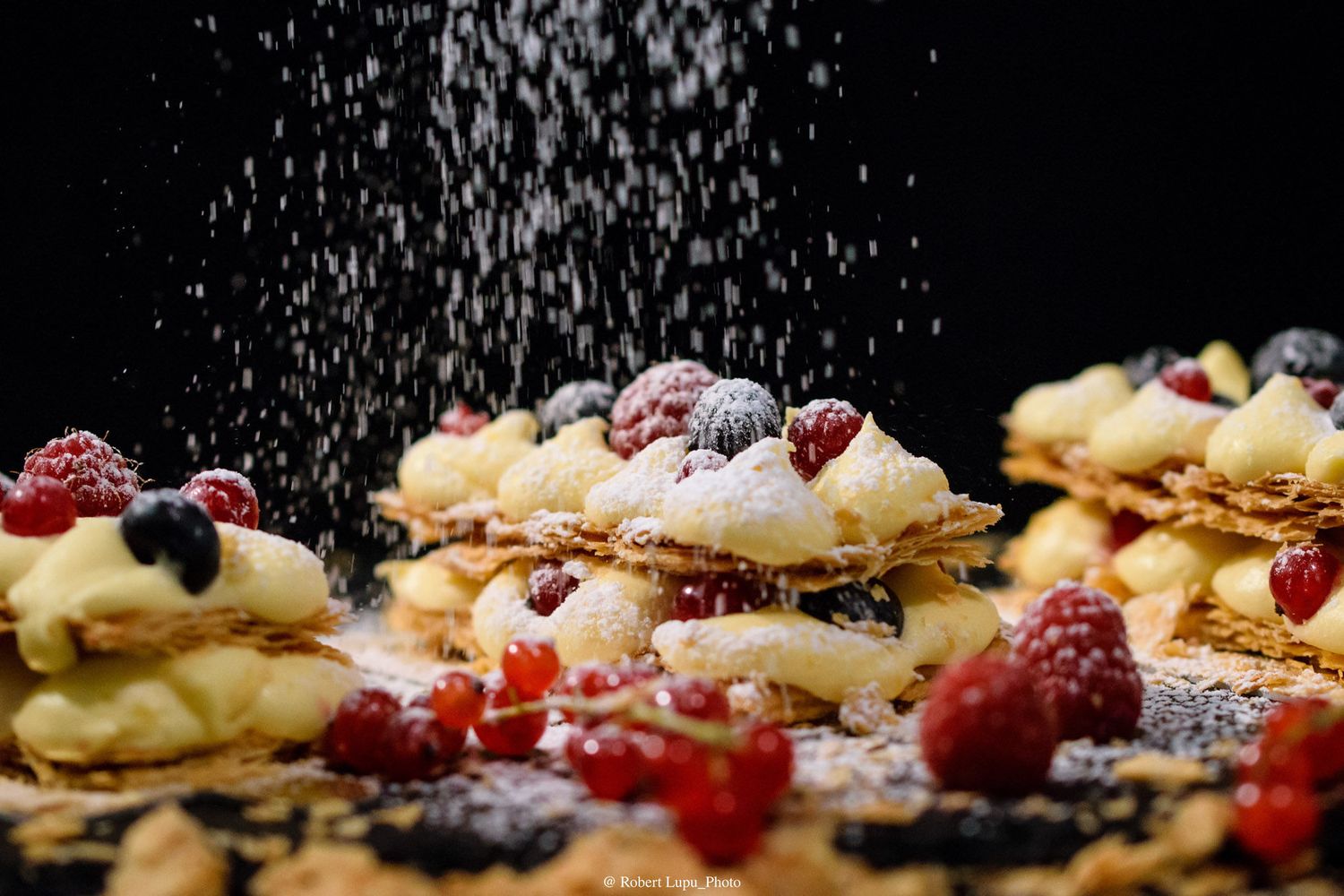 Powdered sugar being dusted over layered pastries topped with fresh berries, cream, and fruit on a dark background.