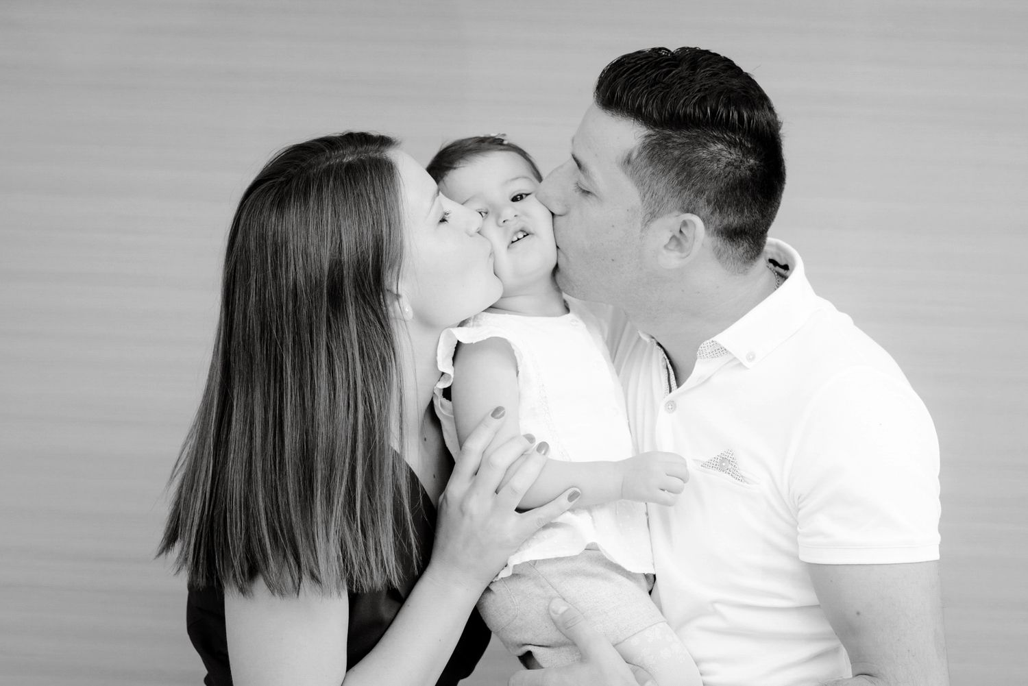 Black and white photo of parents kissing their baby on the cheeks in a studio portrait.