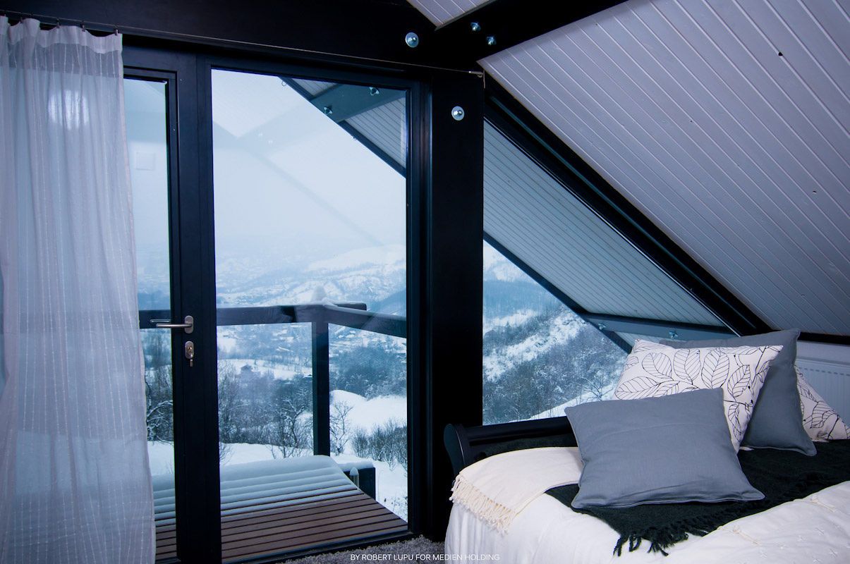 Cozy bedroom with panoramic snowy mountain view through large black-framed glass doors in winter chalet.