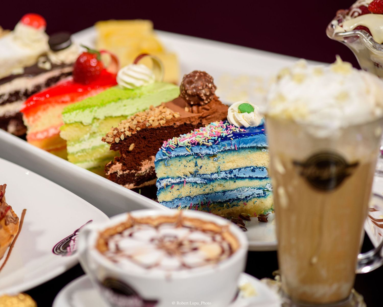 Assorted colorful cake slices and creamy coffee drinks displayed on a table at a dessert cafe.