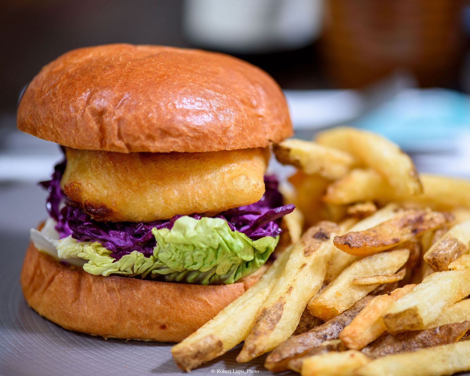 A golden brioche burger with crispy onion ring, red cabbage, and lettuce served with thick-cut fries on a plate.