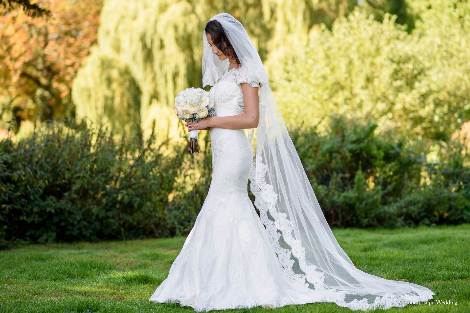 Bride in white mermaid gown with lace veil holding bouquet, standing in lush green garden on wedding day.