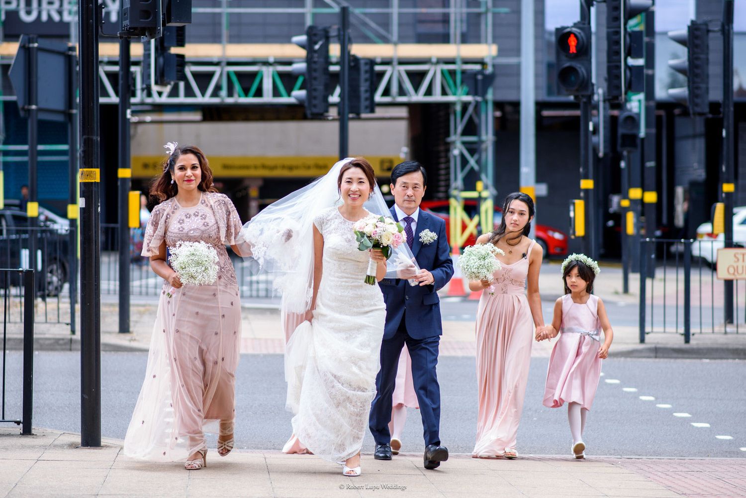 Bride and groom walking with bridesmaids and flower girl at urban crosswalk on wedding day.